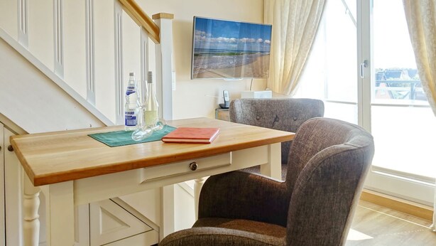 Ein Zimmer mit Schreibtisch aus Holz, zwei Stühlen, einem Fenster mit Vorhängen, einem Gemälde und einer Wasserflasche.
