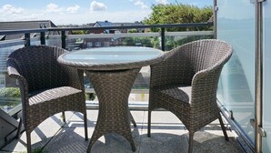 Un balcon doté de fauteuils en osier et d’une table en verre, offrant une vue sur un quartier résidentiel et une étendue d’eau.