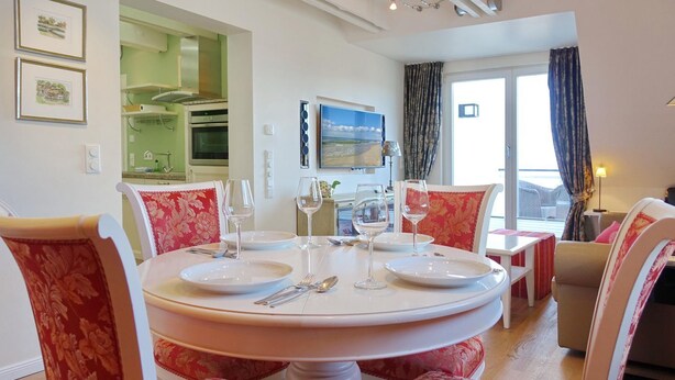 A dining area with a round table set for four, surrounded by floral-patterned chairs. A kitchen with green cabinets and a built-in oven is visible in the background. - Haus Silvana, App. 6 - 2-raum Appartement mit ca. 50m ² Nutzfläche im Obergeschoss (Wenningstedt-Braderup (Sylt))