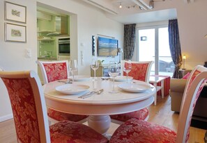 A dining area with a round table set for four, surrounded by floral-patterned chairs. A kitchen with green cabinets and a built-in oven is visible in the background.