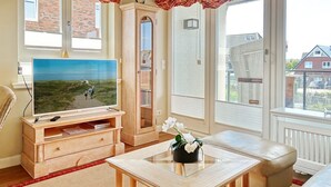 Ein Wohnzimmer mit Fernseher, Holzmöbeln, einem Glastisch und Blick auf ein Backsteinhaus.