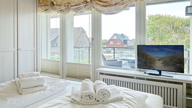 Ein Schlafzimmer mit Bett, Fernseher und Blick auf Häuser durch große Fenster.