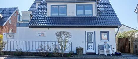Een woonhuis met twee verdiepingen, een pannendak, een voordeur en een klein portaal met een bankje.