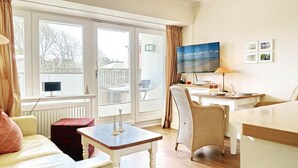 Een gezellige woonkamer met een bank, een kleine tafel, een rieten stoel, een televisie en een eettafel met stoelen.