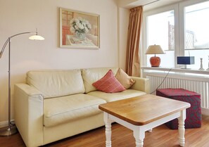 Una sala de estar con un sofá color beige, una mesa de centro de madera, una lámpara de pie, un cuadro, una ventana con cortinas, una lámpara, una computadora y un reposapiés estampado.