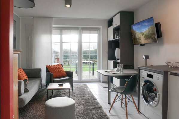 A modern living room with a sofa, armchair, and ottoman, a dining area with a table and chairs, and a kitchen with a washing machine. - Am Kurpark, App. 3 - 2-raum-appartement im Erd- und UG mit ca. 53m² Nutzfläche für Max. Zwei (Wenningstedt-Braderup (Sylt))