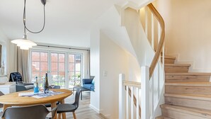 A modern living room with a wooden staircase, a dining area with a round table, and a view of the outdoors through large windows. - Fitte Huck - Hausteil mit ca. 95qm Nutzfläche im EG, UG, OG und DG (Hörnum (Sylt))