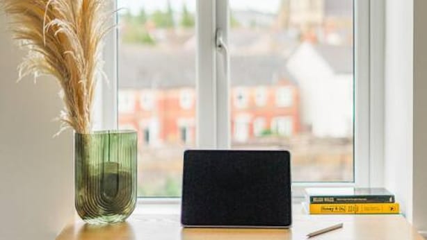 Un bureau avec un ordinateur portable, un vase contenant des plantes séchées et des livres, placé devant une fenêtre donnant sur des immeubles.