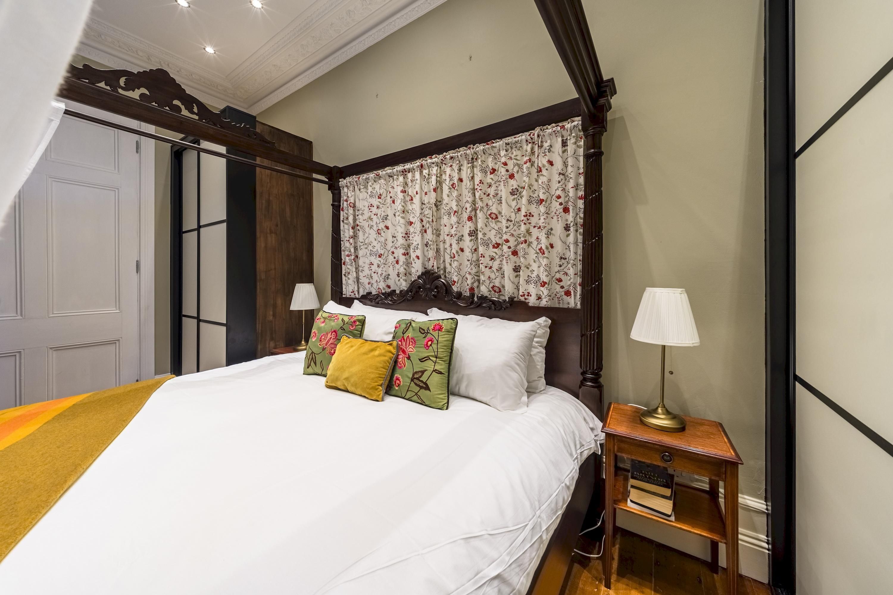 A bedroom with a four-poster bed, floral curtains, and a bedside table with a lamp and books.