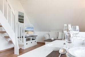 Een moderne, witte woonkamer met een televisie, een bank en een eethoek met een tafel gedekt voor twee personen.