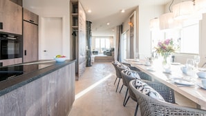 Une cuisine moderne dotée d’un îlot central, d’appareils électroménagers intégrés et d’un coin repas avec des chaises en osier et une longue table.