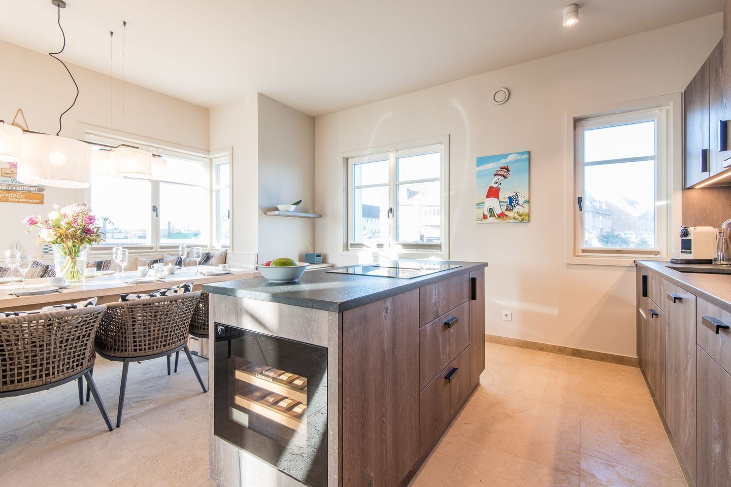 Una cocina moderna con una isla central, comedor con sillas de mimbre y ventanas amplias que dejan entrar luz natural.