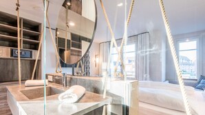 Une chambre d’hôtel moderne dotée d’une baignoire en verre, d’un lit offrant une vue sur la ville et d’un grand miroir.