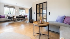 A modern living room with a sofa, a round wooden table with a candle, and a dining area with chairs and a table. - Strandhaus - Hausteil im Eg,ug,og,dg ca. 157qm Nutzfl., Nichtraucher, Haustiere Nicht Erlaubt (Wenningstedt-Braderup (Sylt))