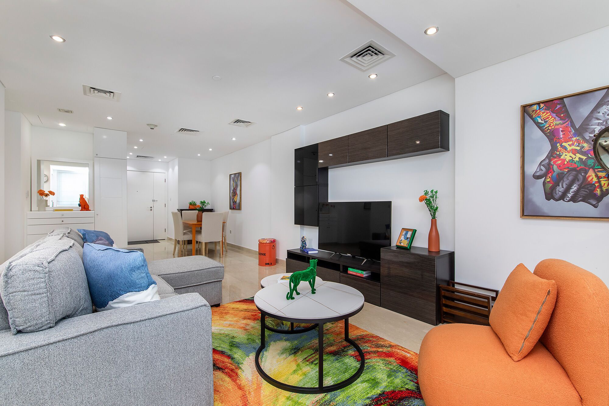 A modern living room with a sofa, a coffee table, and a TV. There is a dining area with a table and chairs, and a kitchen area with cabinets and a microwave.
