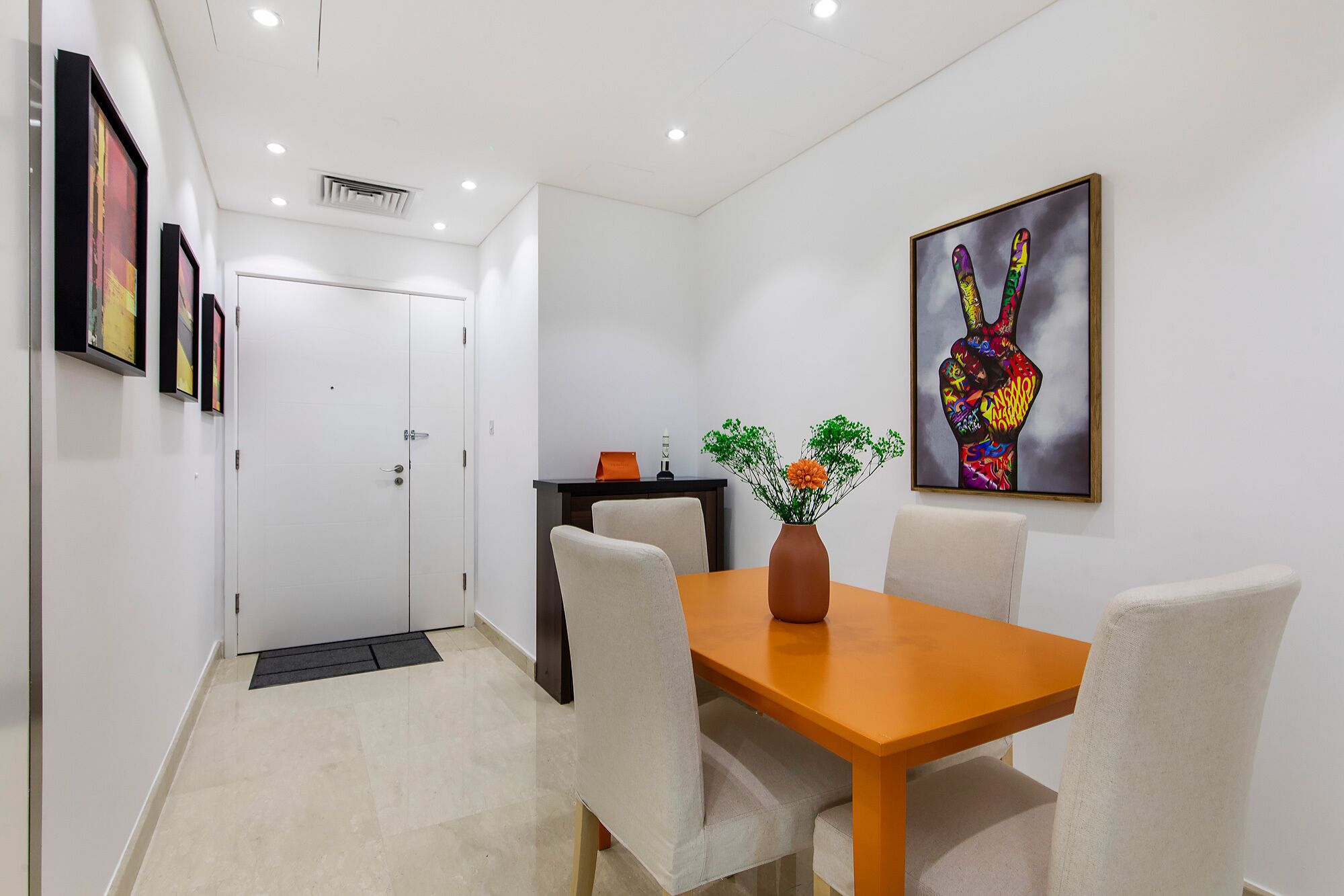 Una habitación de hotel moderna con comedor que incluye una mesa de madera y sillas blancas, un jarrón con flores y una obra de arte colorida en la pared.