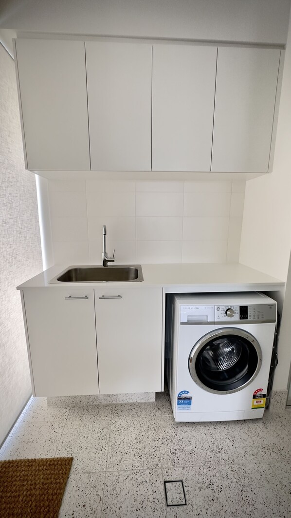 A compact kitchen with a built-in washing machine, a sink, and cabinets. - GRACE - Timeless Style in Central Location (West Busselton)