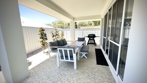 En overdækket terrasse med bord og stole, grill og udsigt til baghaven.