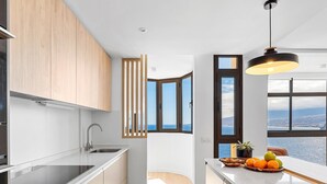 A modern kitchen with a large window, a view of the sea, and a fruit bowl on the counter.