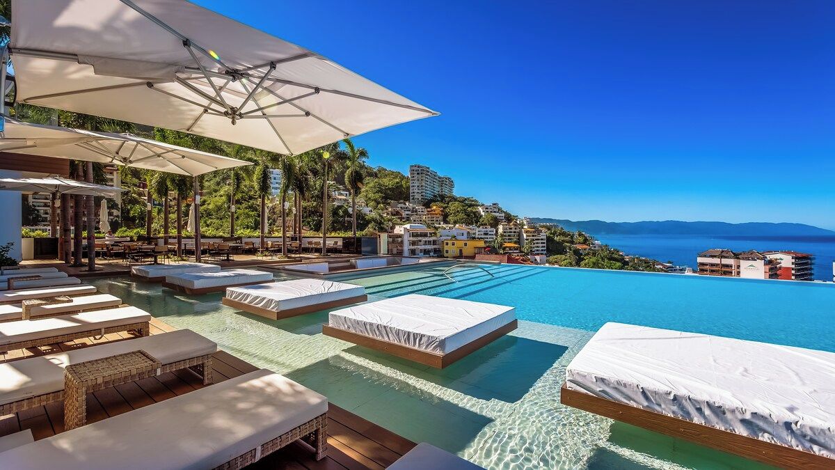 A poolside area with sun loungers and umbrellas, a clear view of the sea, and a hillside with buildings.
