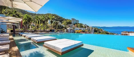 A poolside area with lounge chairs and a bed, overlooking a clear blue sea and a hillside with buildings.