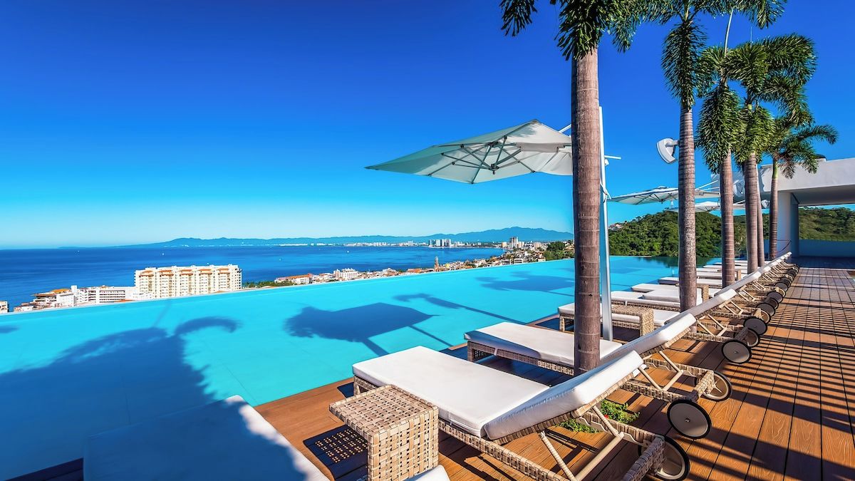 A poolside area with lounge chairs and umbrellas, overlooking a clear blue sea and distant cityscape.