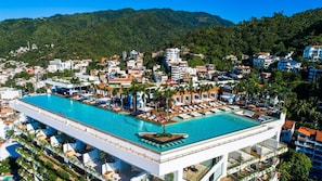 A hotel with a rooftop infinity pool, surrounded by palm trees and a view of a mountainous landscape.