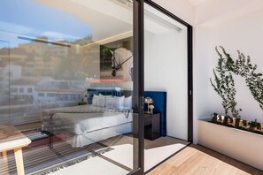 Un dormitorio moderno con puerta de vidrio que da a un balcón, cama con ropa blanca, mesita de noche pequeña y una planta decorativa.