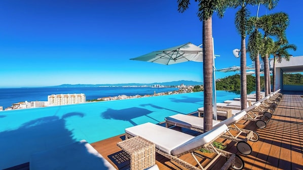 A poolside area with lounge chairs and umbrellas, overlooking a clear blue sea and distant cityscape.