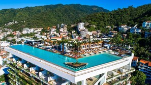 A hotel with a rooftop infinity pool, surrounded by palm trees and a view of a mountainous landscape.