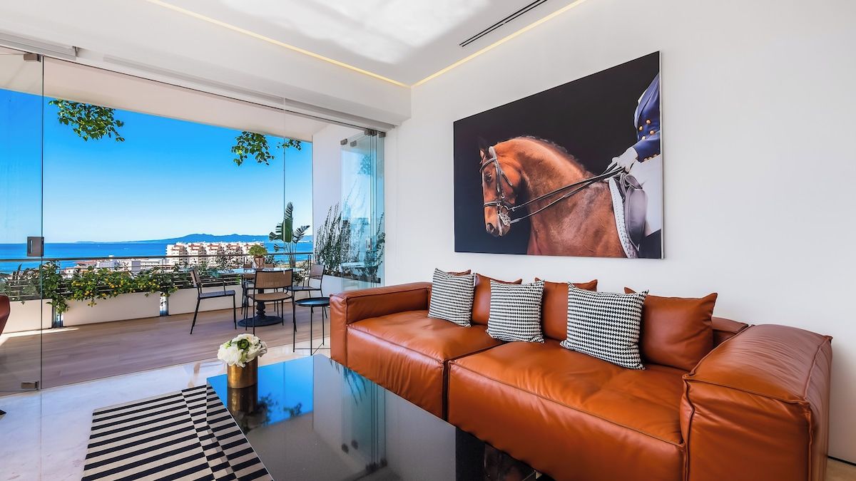 A modern living room with a leather sofa, a glass coffee table, and a large window overlooking a coastal view.