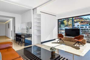 A modern living room with a glass coffee table, a sofa, and a TV. There is a city view through large windows.