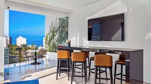 A modern balcony with a glass floor, a dining area with bar stools, a flat-screen TV mounted on the wall, and a view of the ocean and cityscape.