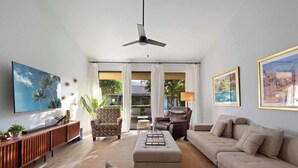 Una sala de estar moderna con un sofá, un sillón, un reposapiés y un ventilador de techo. Hay un televisor montado en la pared y una ventana con cortinas.