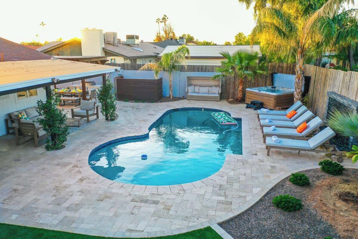 Un patio trasero con piscina, sillas de descanso, jacuzzi y un porche cubierto.