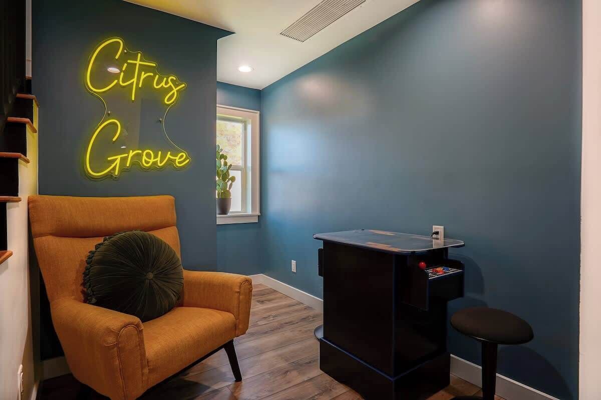 A room with a neon sign reading 'Citrus Grove,' a wooden chair, a small black table, and a window with a plant.