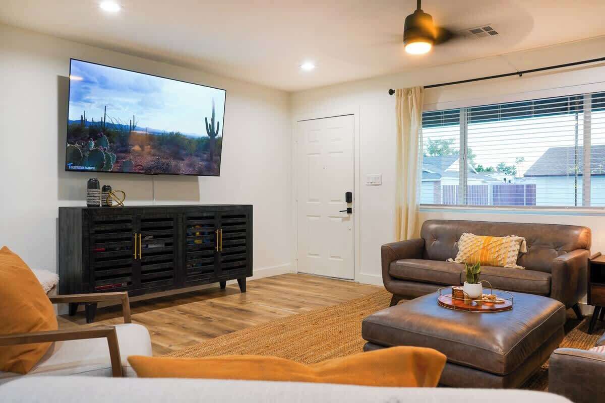 A living room with a flat-screen TV, a dark wooden cabinet, a brown leather sofa, and a window with blinds.