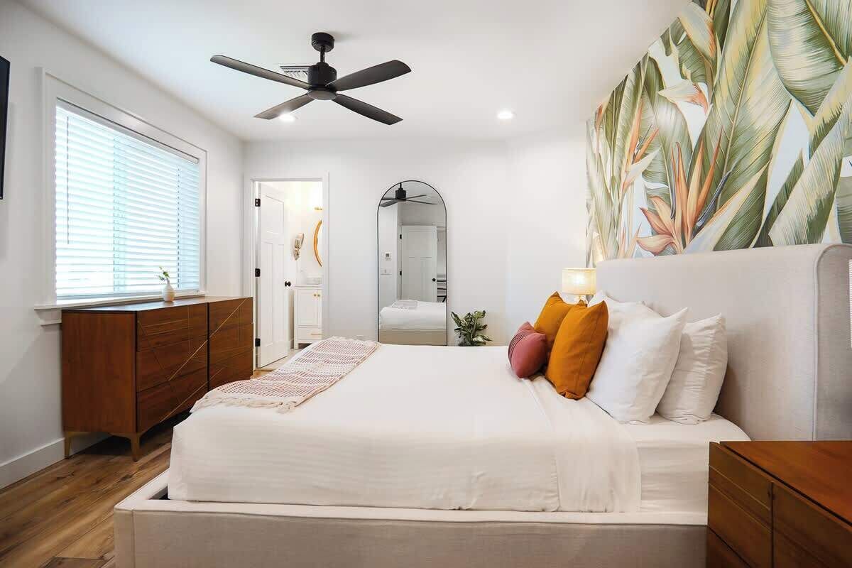 Un dormitorio con cama, ventilador de techo, espejo grande y pared con estampado de hojas tropicales.