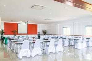 Un sal ón de conferencias con filas de mesas y sillas, una pantalla de proyector y una pared roja de acento.