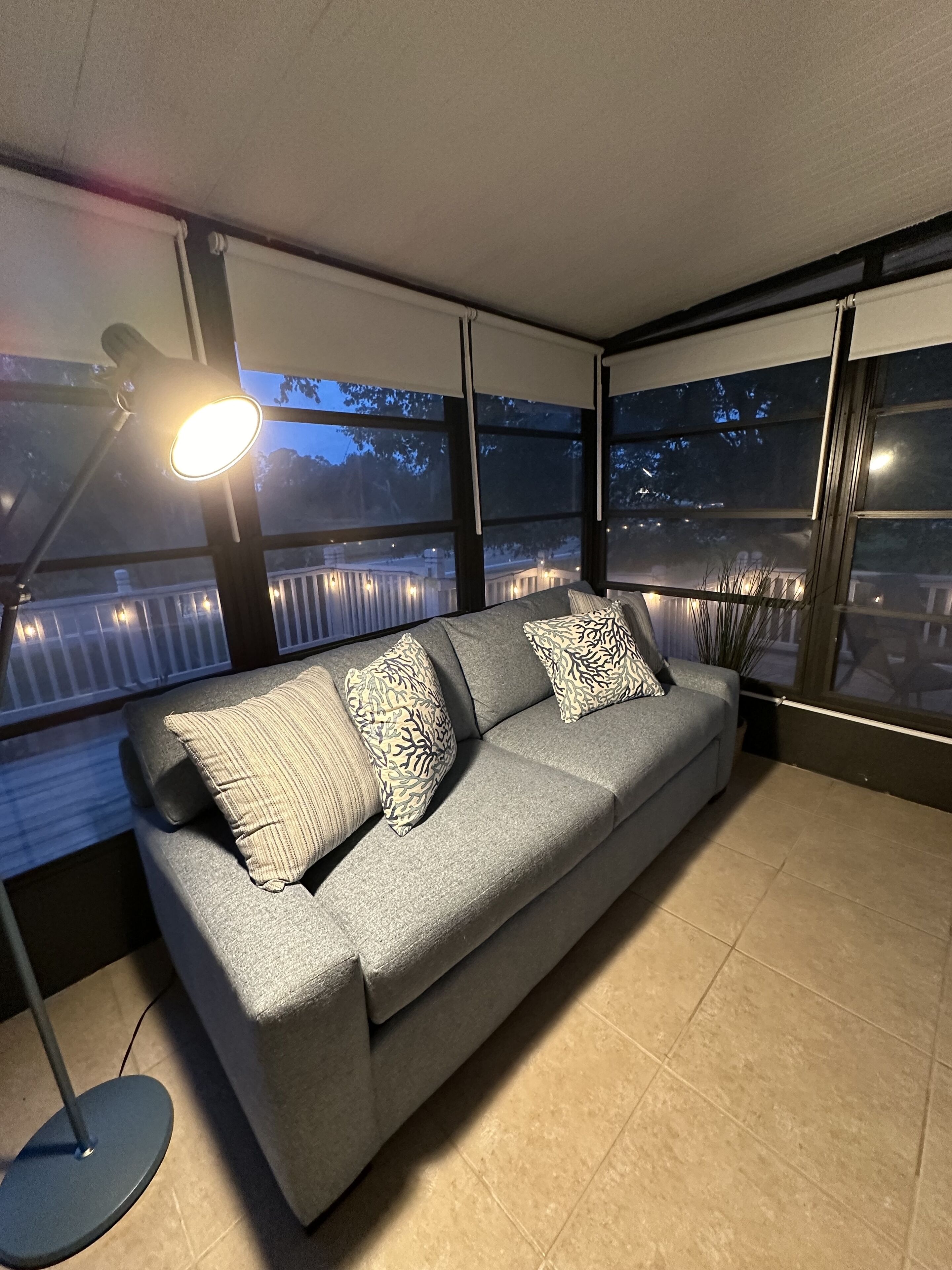 Een grijze bank met gekleurde kussens, een vloerlamp en grote ramen met uitzicht op een balkon en bomen.