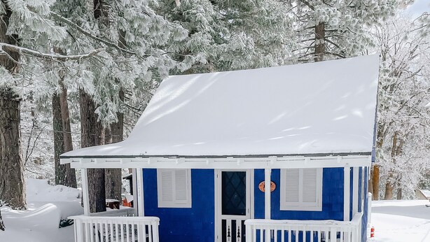 Una cabaña pequeña cubierta de nieve con un porche blanco y fachada azul.