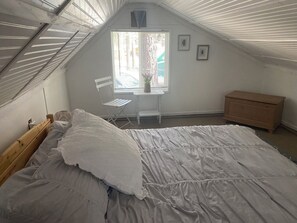 A bedroom with a sloped ceiling, a bed, a small table with a plant, a chair, and a dresser.
