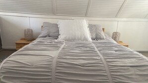 Una cama con edredón blanco, almohadas grises, mesitas de noche de madera y techo blanco.