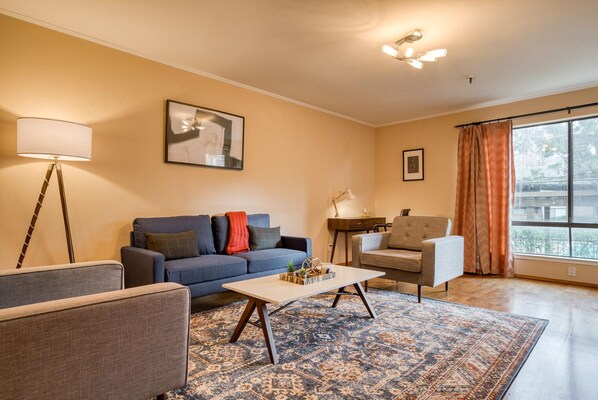 Cozy living room with a comfortable sofa, armchair, and side table, surrounded by artwork and a large window.