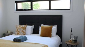 A neatly made bed with a headboard, a small bedside table with a lamp, and a window above the bed. - CBD Elizabeth St Modern Townhouse - Secure Parking (Launceston)