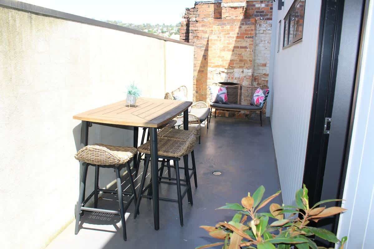 Een balkon met een houten tafel en rieten stoelen, een bakstenen muur en een deur.