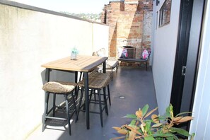 Ein Balkon mit Holztisch und Korbstühlen, einer Ziegelwand und einer Tür.