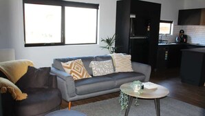 A modern living room with a grey sofa, a round coffee table, and a small ottoman. A kitchen area is visible in the background with a black refrigerator and a window. - CBD Elizabeth St Modern Townhouse - Secure Parking (Launceston)