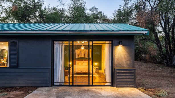 A small, dark-colored building with a green roof and a sliding glass door leading to a bedroom with a bed and bedside lamps. - Manzanita Rest (Mariposa County)