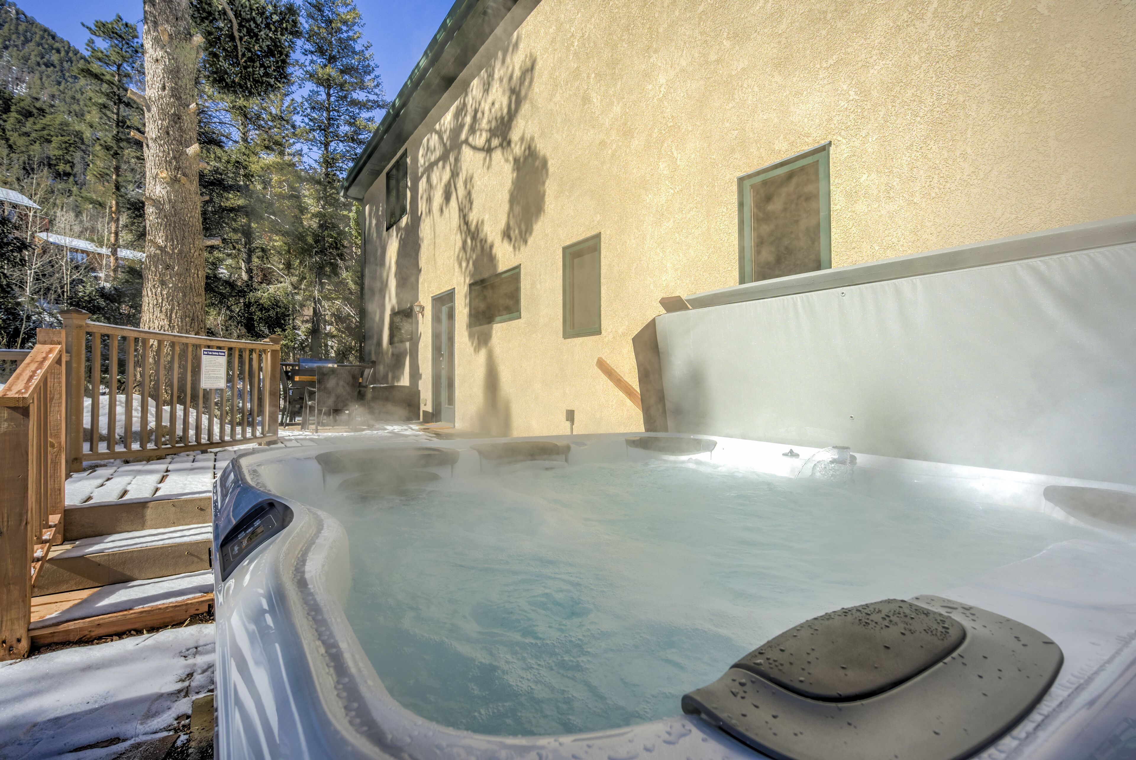 A hot tub area with a wooden deck and steps leading up to it, surrounded by trees and a building with a beige exterior.